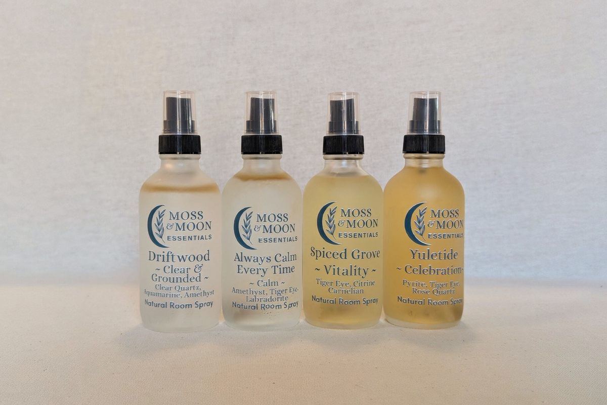 Four Moos & Moon Essences natural room sprays in clear bottles with black spray tops, arranged in a row on a light beige textured background. The bottles contain liquids of varying translucent colors and feature labels for Driftwood, Always Calm, Spiced Rose, and Yuleide.