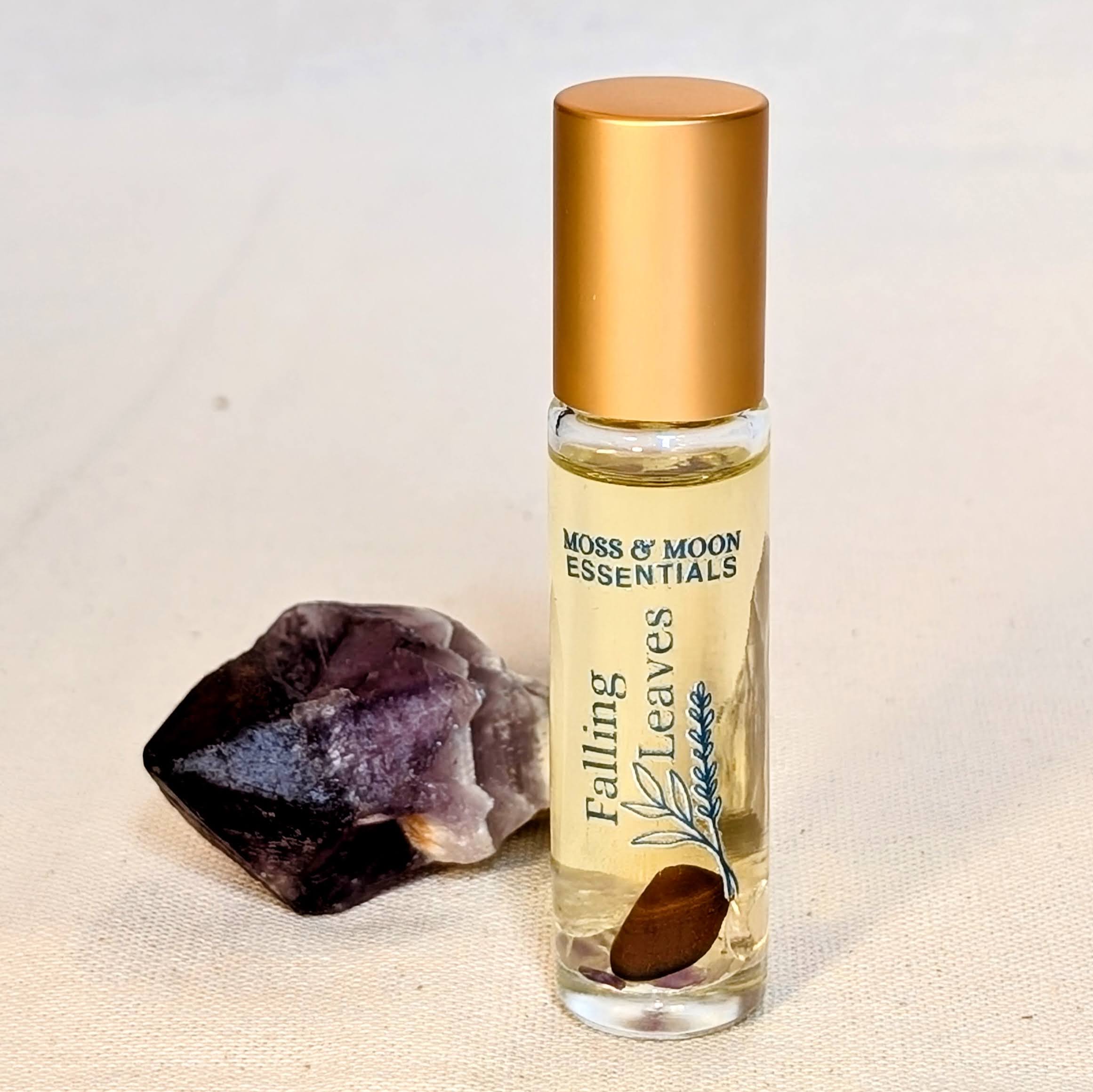 Roll-on bottle of 'Falling Leaves' essential oil by Moss & Moon Essentials with an amethyst crystal on a beige background.